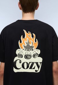 Mustapaitainen puuvillapaita, jossa on graafinen kuva nuotiosta oransseine lieskoineen ja sana "Cozy" lihavoidulla, kermankeltaisella tekstillä alla.