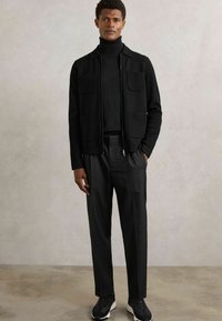 Juodas zip-up džemperis su dviem priekinėmis kišenėmis, uždėtas ant juodos golfo megztinio, derinamas su juodomis oficialiomis kelnėmis ir juodais sportbačiais.