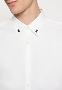 Chemise blanche avec col pointu, accents métalliques ornés de motifs noirs, et boutons nacrés sur le devant.