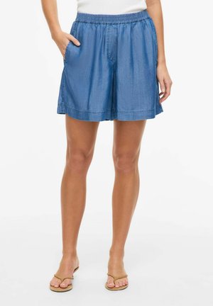 Femeie purtând pantaloni scurți din denim albastru largi, cu talie elastică și buzunare laterale, asortați cu șlapi bej pe fundal alb.