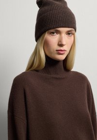 Vrouw met lang blond haar, die een donkerbruin gebreid mutsje en een bijpassende coltrui draagt, tegen een effen lichte achtergrond.