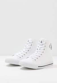 Weiße High-Top-Sneaker aus Canvas mit Metallösen, flachen weißen Schnürsenkeln, einer Gummisohle und einem schwarzen Streifen als Akzent auf der Sohle.