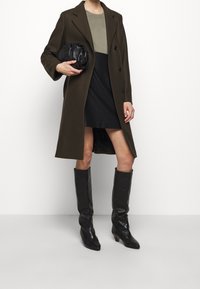 Olivgrünener Wollmantel mit tailliertem Design, schwarzer Minirock und hohen schwarzen Lederstiefeln, kombiniert mit einer schwarzen Clutch.