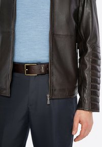Giacca in pelle marrone con dettagli a coste sulle maniche, abbinata a un maglione azzurro chiaro, pantaloni scuri e una cintura marrone con fibbia in metallo.