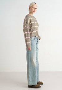 Grijze gebreide trui met golvende roze strepen, gecombineerd met licht gewassen wijde jeans en bruine chunky schoenen, staand tegen een neutrale achtergrond.