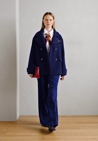 Abrigo de lana azul marino de doble botonadura con grandes solapas, combinado con pantalones de pierna ancha a juego y una camisa blanca, acentuado por una corbata de seda con estampado.