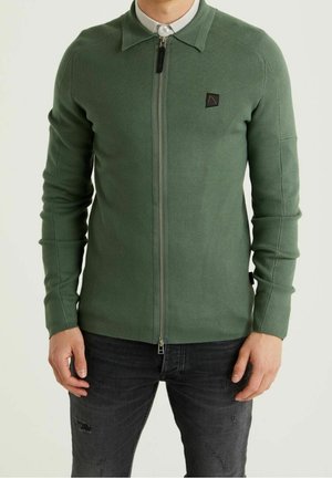 Maglione verde con zip e collo a camicia, texture a coste e una piccola patch nera con logo sul petto. Indossato sopra jeans scuri.