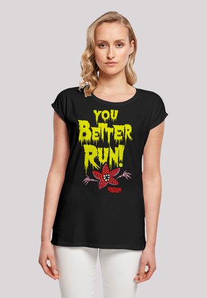 Schwarzes T-Shirt mit kurzen Ärmeln, das mit leuchtend gelbem und rotem Grafiktext und Design ausgestattet ist, sowie einem Blumenmotiv und Branding von "Stranger Things".