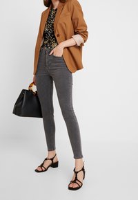 Blazer marrón sobre blusa negra con flores, jeans skinny grises, bolso negro con asa de carey y sandalias negras de tacón cuadrado.