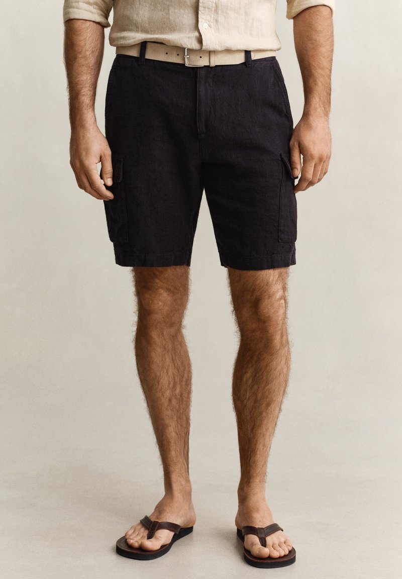 Homme portant une chemise beige à manches longues, un short cargo noir avec des poches à rabat, une ceinture beige et des sandales marron foncé, debout sur un fond uni.