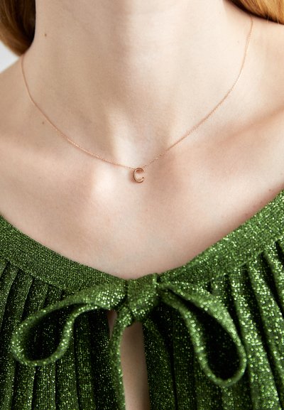 Collier en or rose avec un petit pendentif en forme de lettre 'C' sur une chaîne délicate, porté contre un haut plissé vert pailleté avec un détail en nœud.