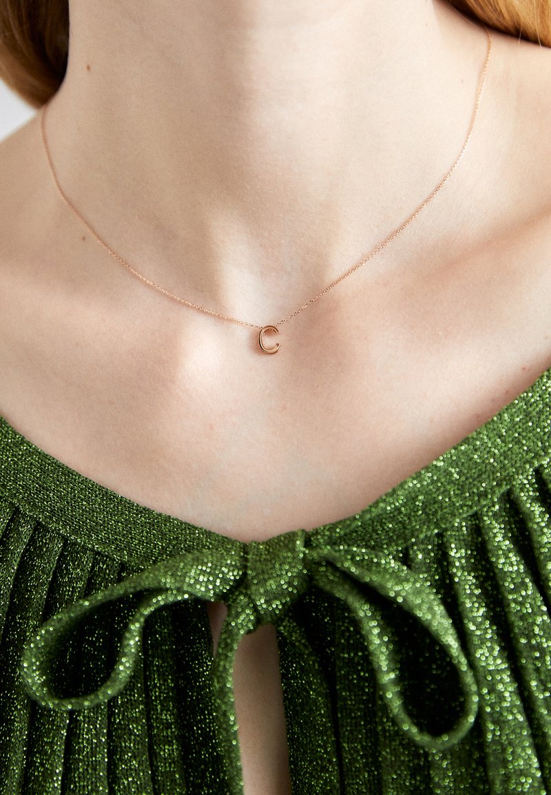 Rožu zeltījuma kaklarota ar mazu burtu 'C' kulonu uz delikāta ķēdes, uzvilkta pret zaļu, spīdīgu, krokotu topu ar loku detaļu.