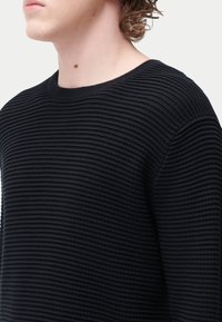 Pull noir texturé avec un design côtelé, un col rond et des manches longues, mettant en valeur un motif horizontal sur l'ensemble.