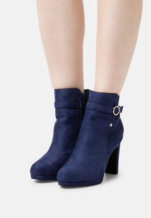 Bottes chevilles en daim bleu marine à talon haut, avec une fermeture éclair latérale et un accent circulaire doré sur la bride. Texture lisse, forme épurée.