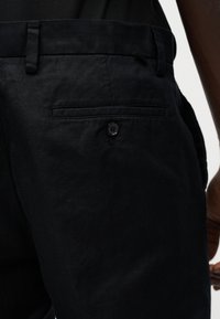 Κοντινή εικόνα μαύρου παντελονιού που δείχνει πίσω τσέπη με κουμπί και θηλιά για ζώνη, μερική άποψη του ισχίου και του χεριού ενός ατόμου.