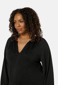 Mustapaita, jossa on V-mallinen kaula-aukko ja nyörity yksityiskohtia. Ominaisuuksiin kuuluu pitkät hihat ja sileä pinta, joka sopii sekä vapaa-aikaan että muodollisiin tilaisuuksiin.