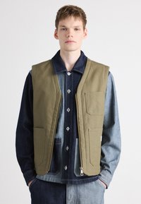 Olive green sleeveless vest with large front pockets, zip closure, and beige accents. Worn over a dark denim shirt with light denim details.