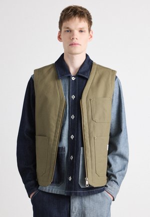 Gilet sans manches vert olive avec de grandes poches avant, fermeture éclair et accents beige. Porté par-dessus une chemise en denim foncé avec des détails en denim clair.