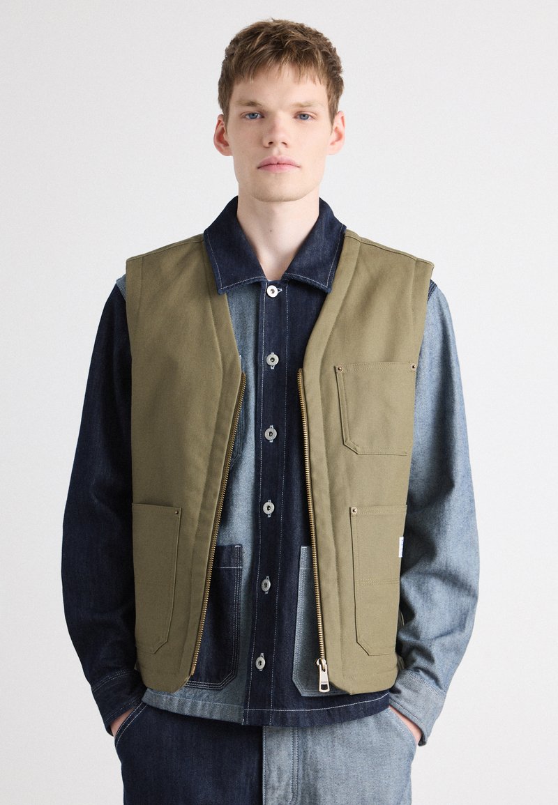 Olive green sleeveless vest with large front pockets, zip closure, and beige accents. Worn over a dark denim shirt with light denim details.