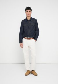 Donkerblauw denim overhemd met twee borstzakken, gecombineerd met witte broek en lichtbruine sneakers. De outfit wordt gecompleteerd met een bruine riem.