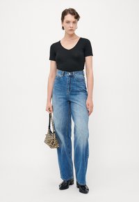 Frau in schwarzem kurzärmligem Oberteil und hochtaillierten blauen Jeans, die eine kleine Handtasche mit Zebramuster hält und vor einem weißen Hintergrund steht.