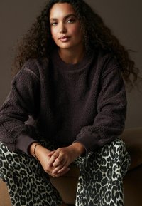 Mörkbrun fleecehoodie med ribbat krage och ärmslut; parat med svarta och vita leopardmönstrade byxor. Modell sitter, händerna hopknäppta.