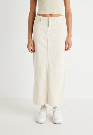 Jupe maxi en denim couleur crème avec une fente à l'avant, taille haute et coutures verticales. Portée avec des baskets blanches pour le contraste.