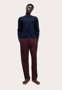 Chemise à manches longues bleu marine, coupe décontractée. Pantalon bordeaux à motifs avec taille élastique. Modèle pieds nus debout devant un fond neutre.