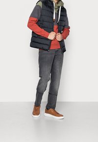 Chaleco aislante negro sobre una sudadera roja con mangas grises, combinado con pantalones grises y zapatillas marrones. Texturas suaves con colores contrastantes.
