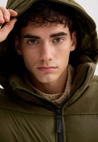 Chaqueta acolchada de color verde oliva con capucha, equipada con cremallera y cuello acanalado. Textura lisa con un suave tejido de punto en el interior. Fondo neutro.