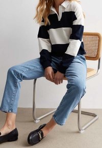 Camisa polo de rayas azul marino y blanca de manga larga, jeans rectos de color azul claro y mocasines negros con herrajes dorados, sentado en una silla de madera.