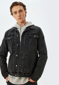 Chaqueta de mezclilla negra con bolsillos en el pecho, botones plateados y un acabado texturizado, superpuesta sobre una sudadera con capucha gris claro.