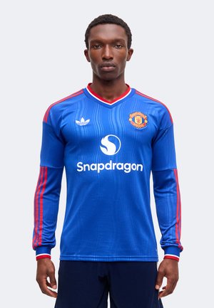 Uomo che indossa una maglia da calcio Manchester United blu a maniche lunghe con strisce rosse, logo Snapdragon e stemma Adidas, in piedi davanti a uno sfondo semplice.