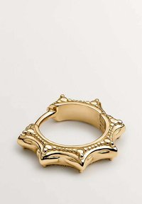 Anillo dorado con puntas redondeadas y detalles de cuentas en un diseño simétrico y ornamentado, mostrado sobre un fondo claro y liso.