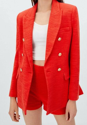Femme portant un blazer rouge texturé à double boutonnage avec des boutons dorés et un short taille haute assorti par-dessus un top court blanc.