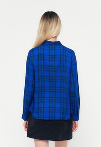 Lange mouwen geruite blouse in blauw met groene en zwarte ruiten. Lichtgewicht stof, knoopmanchetten en licht gebogen zoom.