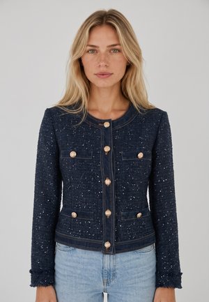 Donna bionda che indossa una giacca blu scuro testurizzata con bottoni dorati e jeans azzurri, in piedi davanti a uno sfondo grigio uniforme.