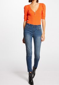 Orangestribet top med korte puffærmer og lynlås fortil, parret med tætsiddende blå denim jeans og sorte ankelstøvler.