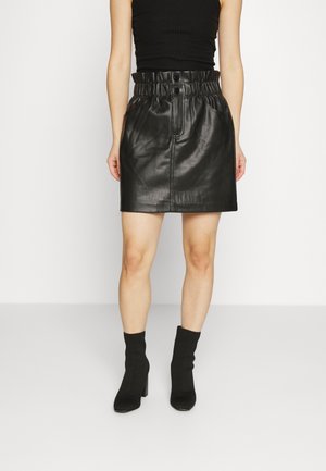 Jupe mini en cuir noir avec une taille froncée, des boutons sur le devant et des poches latérales. Portée avec des bottines noires et un haut noir ajusté.