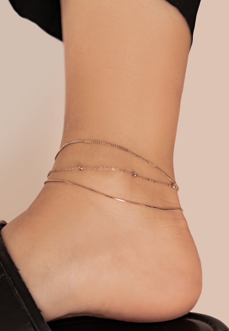 Tobillera de oro que presenta tres hilos: una cadena fina, una delicada cadena de cuentas y una banda pulida y suave. Lleva puesta en un tobillo ligeramente bronceado.