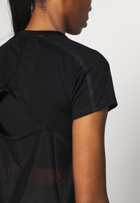 Musta urheilupaita lyhyillä hihoilla, jossa on läpinäkyvä kerros selässä ja kontrastiväriset saumat hartioilla. Sileä rakenne.