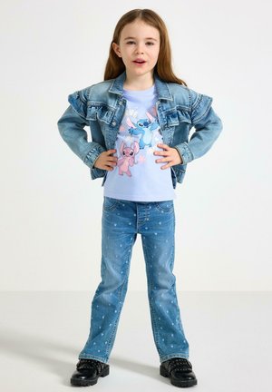 Pige stående med hænderne på hofterne iført en denimjakke, blå t-shirt med grafik, jeans med hjerteprint og sorte støvler med nitter.