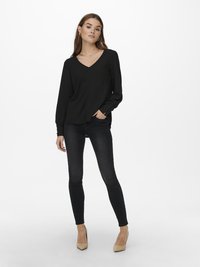 Blusa preta de manga longa com decote em V, tecido texturado e ajuste descontraído, combinada com jeans skinny pretos e saltos bege.