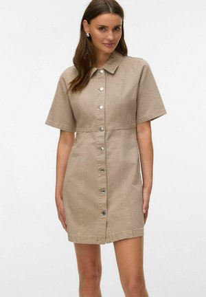 Mujer que lleva un vestido beige de manga corta con botones y cuello, de pie frente a un fondo claro y sencillo.