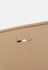 Borsa in pelle beige con texture, dotata di zip e un logo con accenti neri, bianchi e marroni chiari. Superficie liscia con dettagli raffinati.