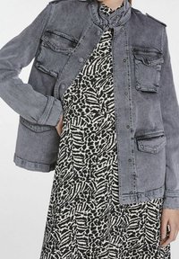Szara kurtka dżinsowa z stójką, zatrzaskami i czterema przednimi kieszeniami, nałożona na sukienkę w czarno-białe abstrakcyjne wzory.