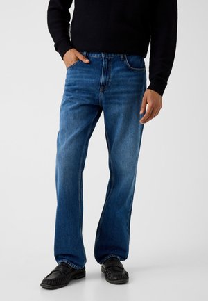 Uomo che indossa jeans blu a gamba dritta, maglione nero e mocassini neri, con una mano in tasca, in piedi su sfondo bianco.