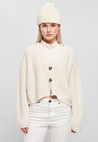 Krämfärgad stickad cardigan med V-kort, oversize-passform, prydd med stora svarta knappar. Bärs över en ljus polotröja och vita cropped byxor.