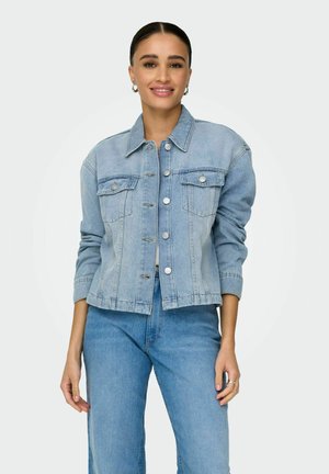 Woman smiling, wearing light blue denim jacket buttoned up and matching blue jeans, standing against plain light background.