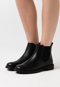 Černé kožené kotníkové boty s elastickými bočními panely, zaoblenou špičkou a strukturovanou gumovou podrážkou. Mají hladký povrch a minimalistický design.